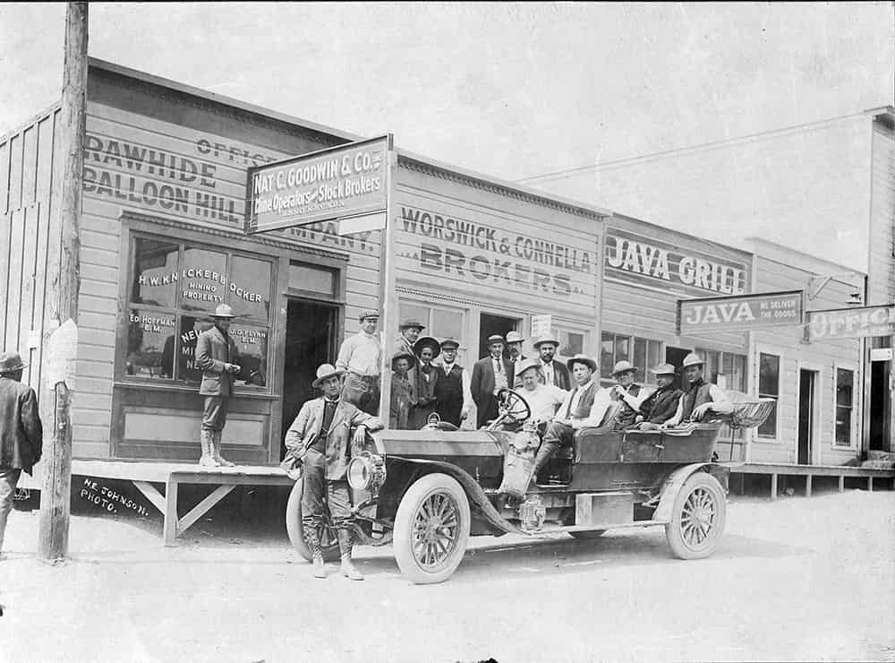 Black and white photograph of a group of men in a car and on the porches of mining stock broker companies in Rawhide